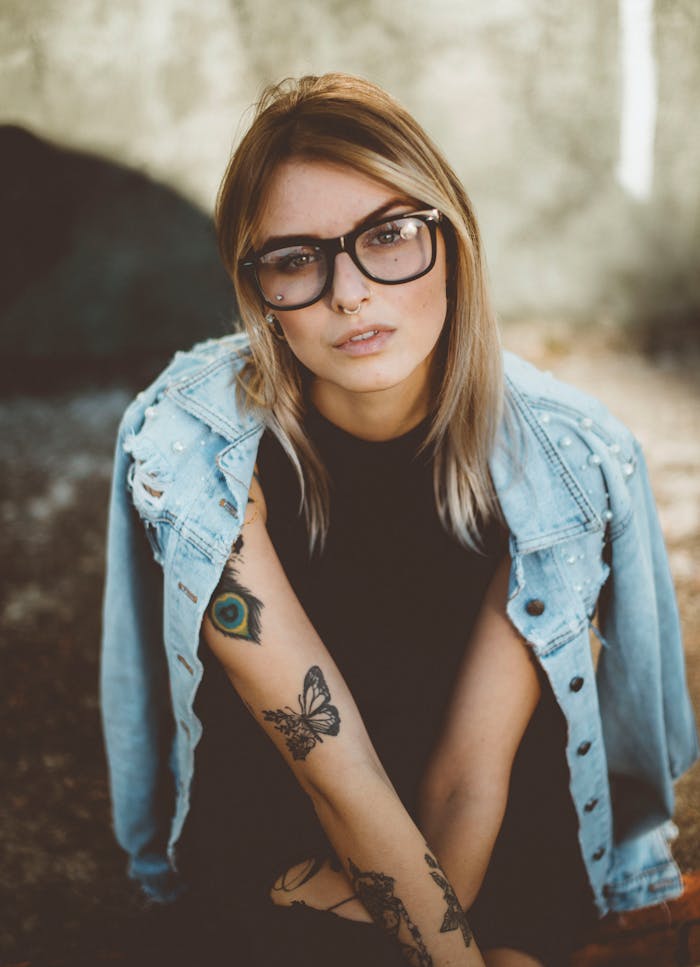 Portrait of a stylish young woman with glasses and tattoos wearing a denim jacket outdoors.