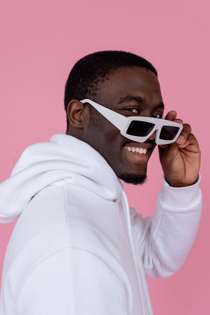 Happy young black guy in white hoodie and sunglasses looking at camera on pink background in bright studio